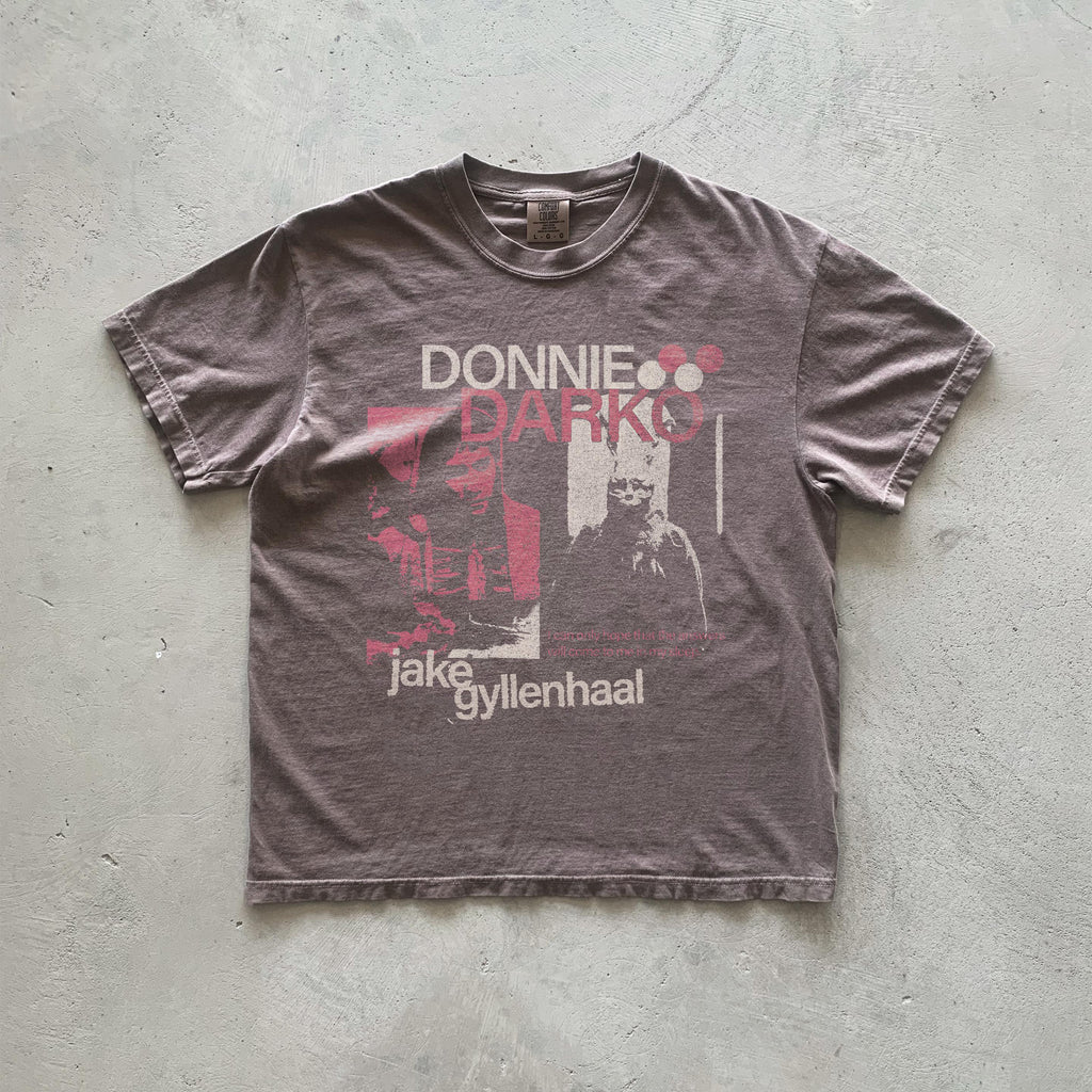 May include: A brown t-shirt with a faded print of the movie Donnie Darko. The print features the title of the movie in red and white text, a black and white image of Jake Gyllenhaal, and the text 'Jake Gyllenhaal' in white.