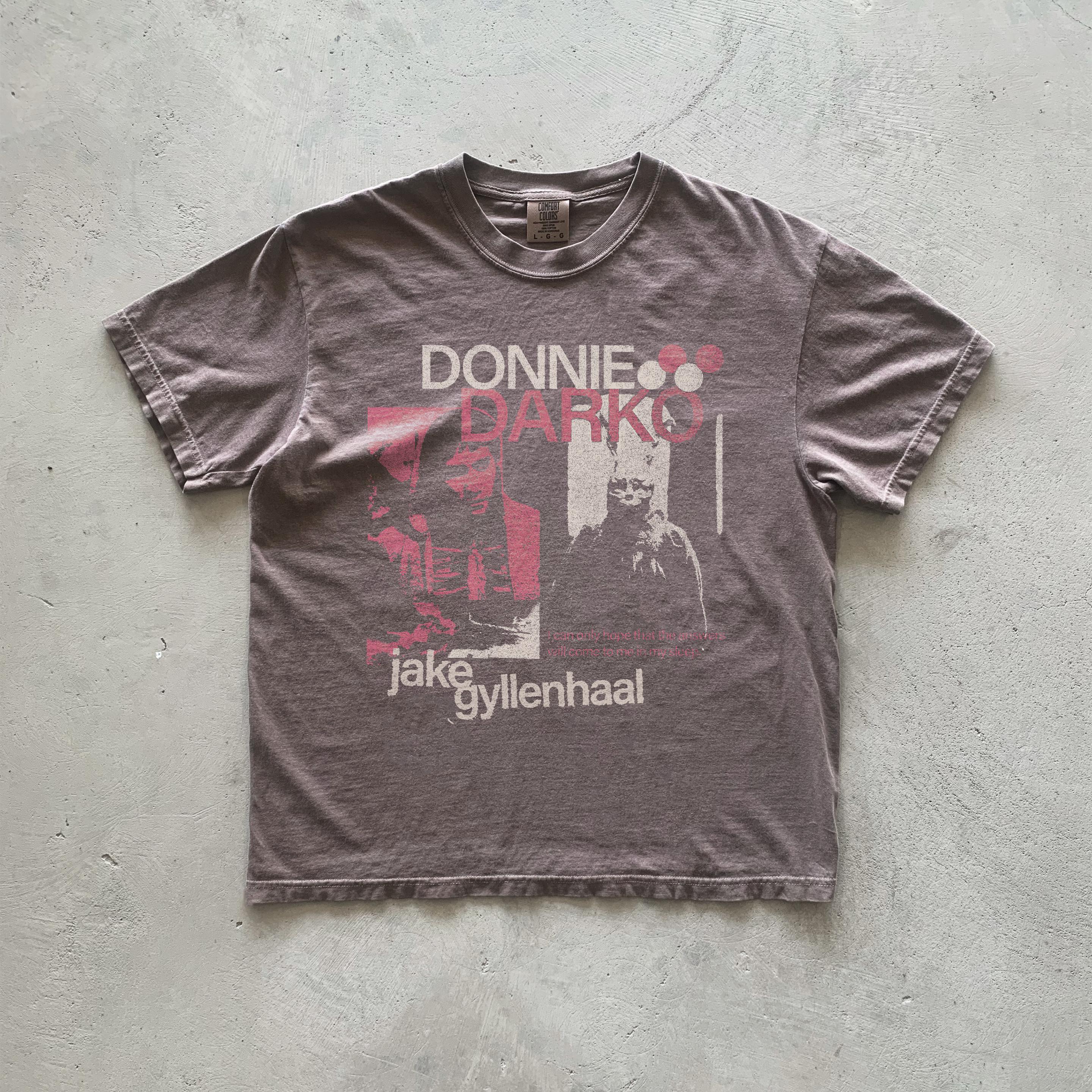 May include: A brown t-shirt with a faded print of the movie Donnie Darko. The print features the title of the movie in red and white text, a black and white image of Jake Gyllenhaal, and the text 'Jake Gyllenhaal' in white.