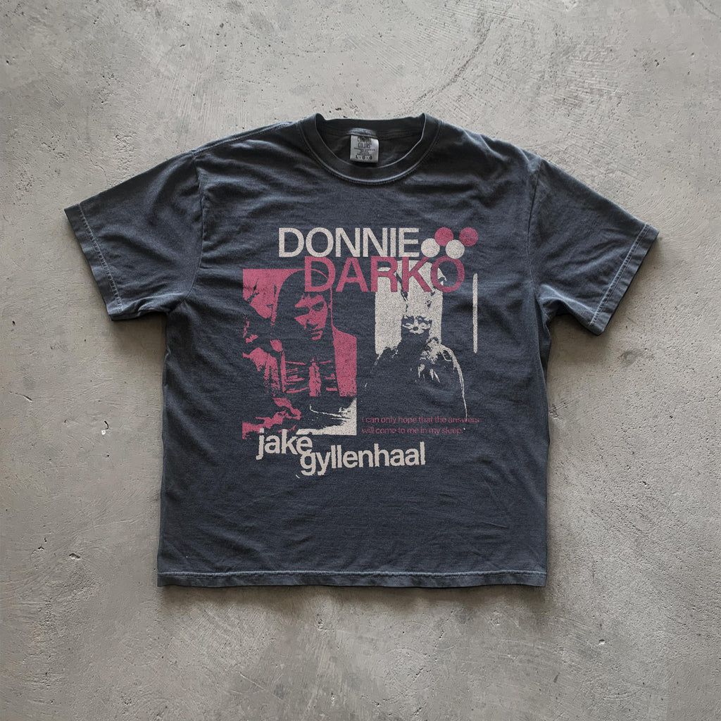May include: A black t-shirt with a white graphic print of Jake Gyllenhaal from the movie Donnie Darko. The graphic includes the movie title 'Donnie Darko' in pink and white text, a quote from the movie, and a black and white image of Jake Gyllenhaal.