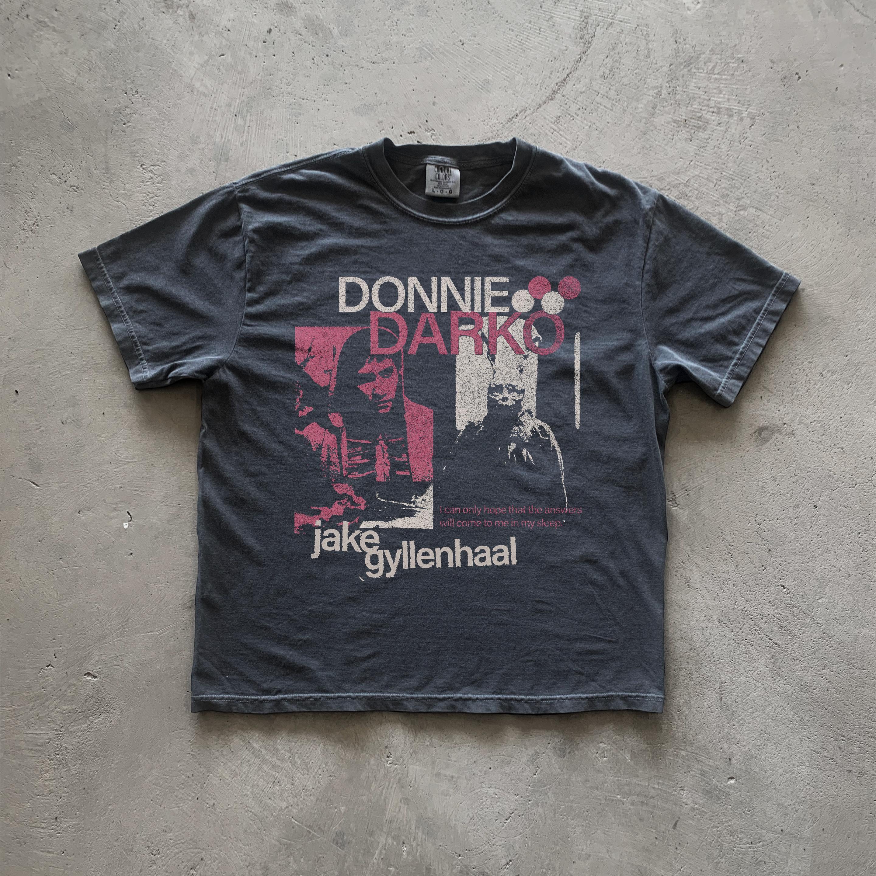 May include: A black t-shirt with a white graphic print of Jake Gyllenhaal from the movie Donnie Darko. The graphic includes the movie title 'Donnie Darko' in pink and white text, a quote from the movie, and a black and white image of Jake Gyllenhaal.
